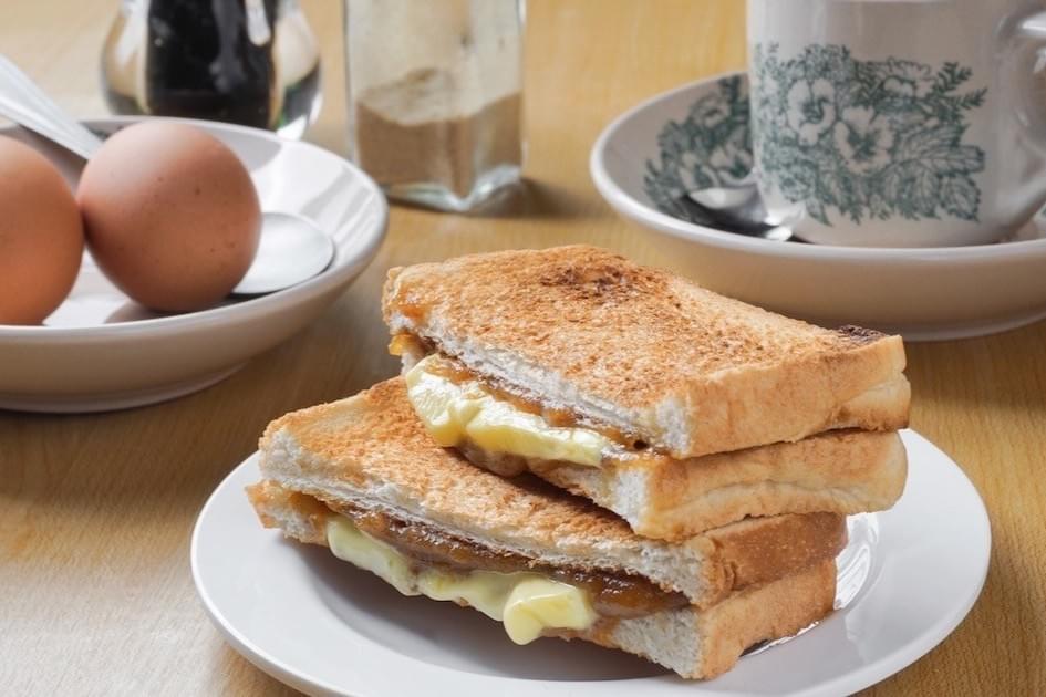This image displays a classic Kaya toast breakfast, featuring crisp, toasted bread filled with thick slabs of butter and coconut jam. The sandwiches are accompanied by a side of soft-boiled eggs and a traditional floral-patterned coffee cup. This image displays a classic Kaya toast breakfast, featuring crisp, toasted bread filled with thick slabs of butter and coconut jam. The sandwiches are accompanied by a side of soft-boiled eggs and a traditional floral-patterned coffee cup.