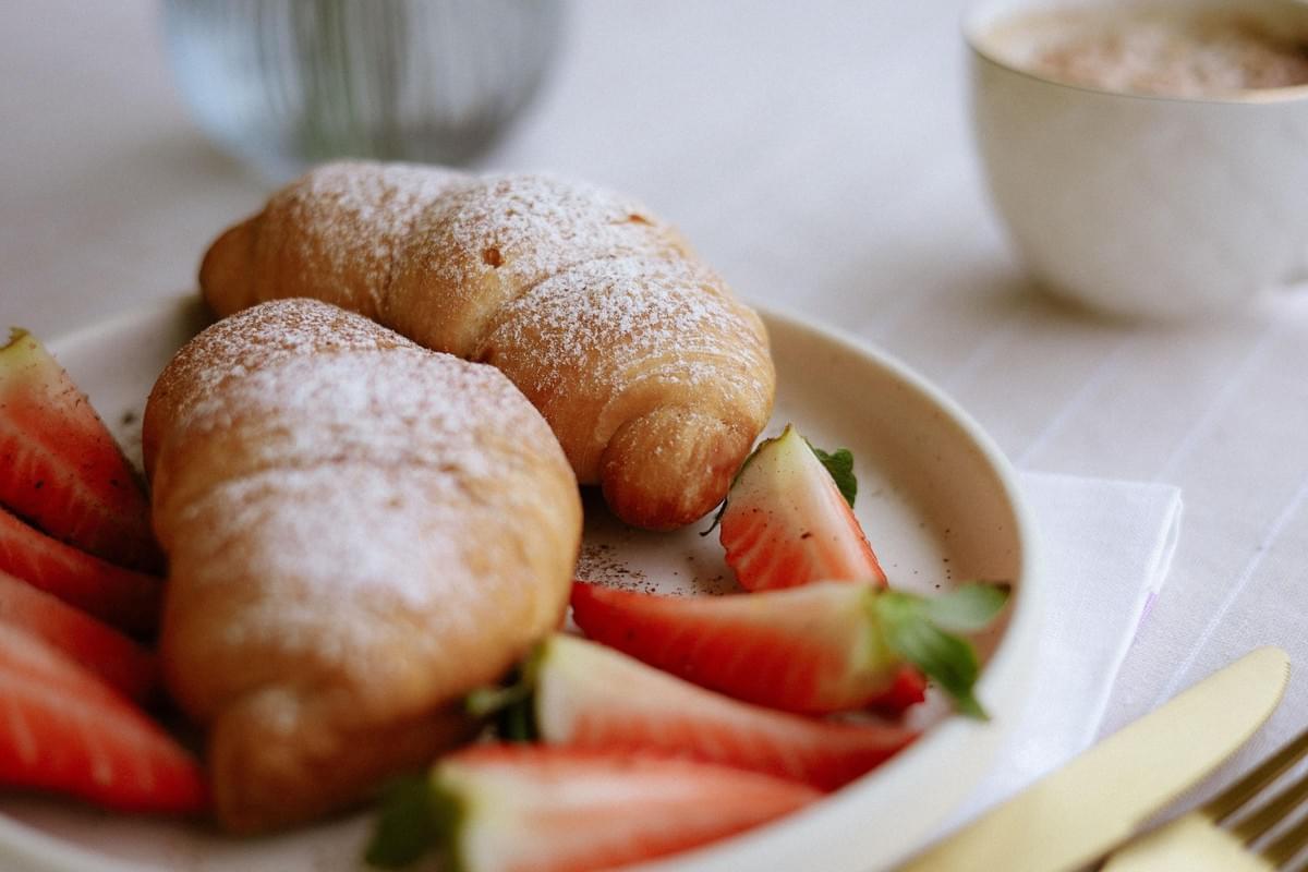 Two golden-brown croissants dusted with powdered sugar sit on a ceramic plate alongside fresh, sliced strawberries. In the softly blurred background, a warm beverage in a white cup and a glass vase rest on a light-colored tablecloth. Two golden-brown croissants dusted with powdered sugar sit on a ceramic plate alongside fresh, sliced strawberries. In the softly blurred background, a warm beverage in a white cup and a glass vase rest on a light-colored tablecloth.