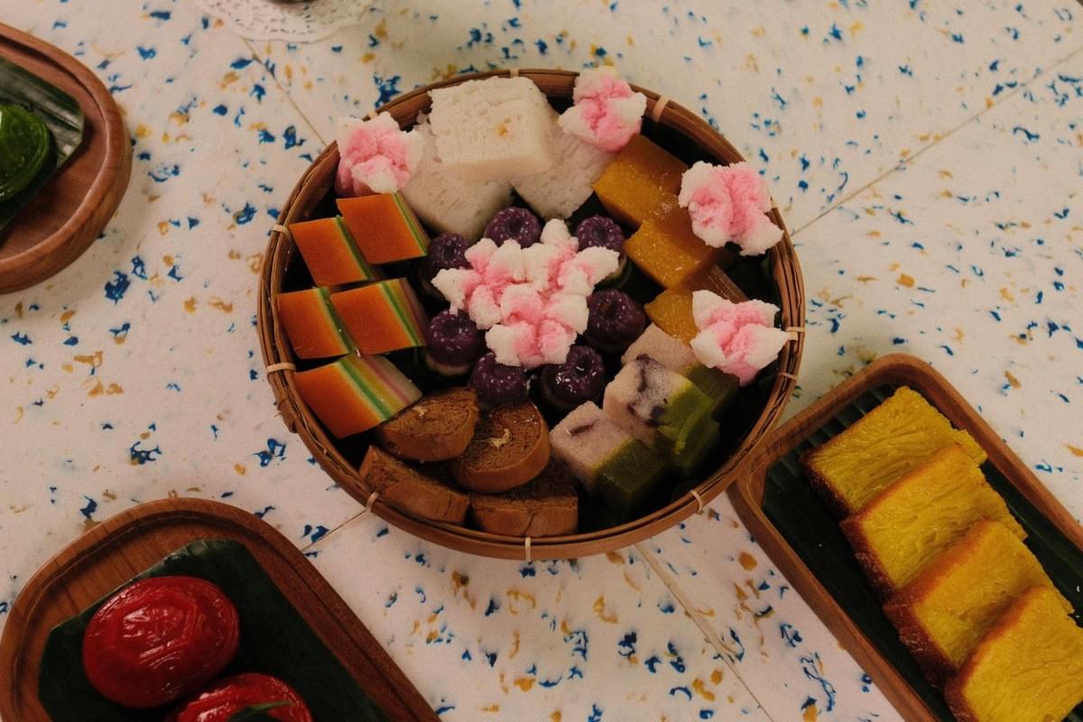 An overhead view shows a round woven basket filled with an assortment of colorful traditional Southeast Asian or Nyonya cakes and steamed sweets. Surrounding the center basket on a speckled tabletop are additional wooden trays displaying slices of yellow cake, red kueh, and other delicacies. An overhead view shows a round woven basket filled with an assortment of colorful traditional Southeast Asian or Nyonya cakes and steamed sweets. Surrounding the center basket on a speckled tabletop are additional wooden trays displaying slices of yellow cake, red kueh, and other delicacies.