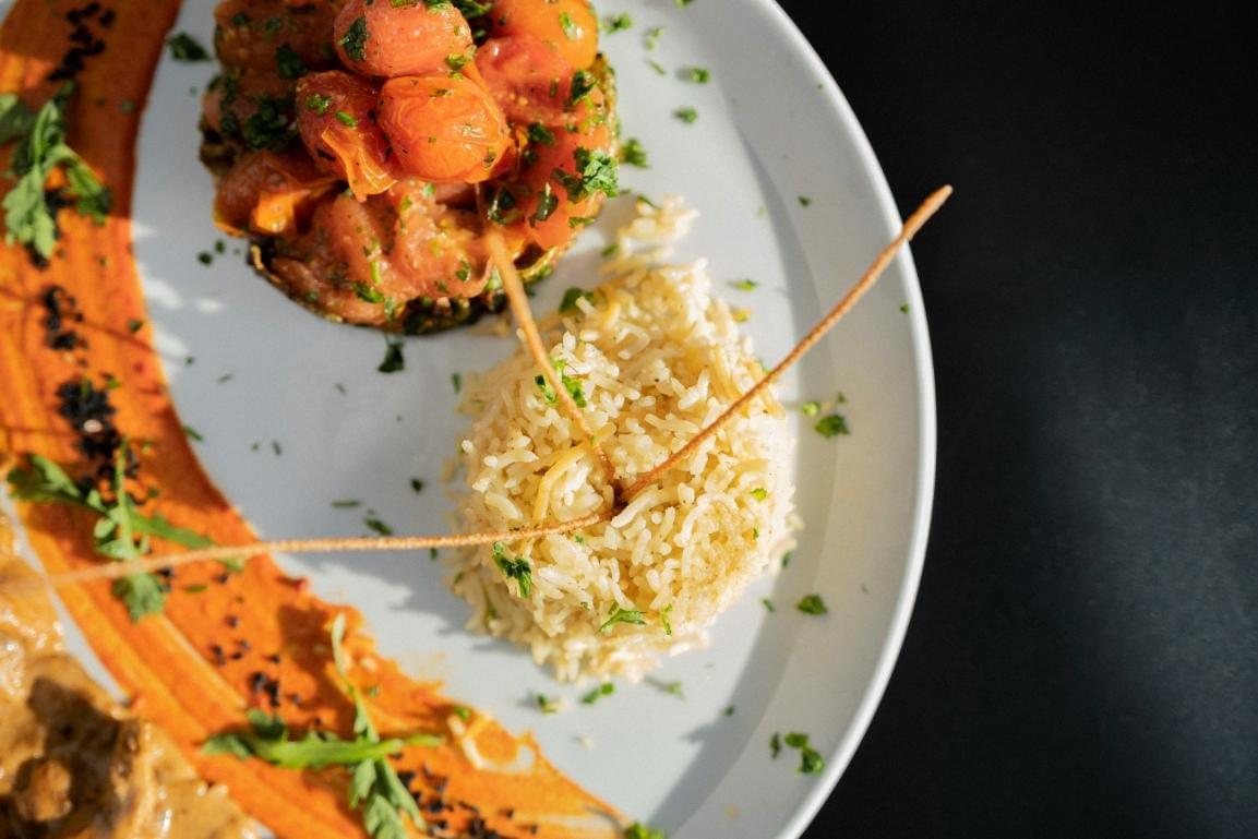 The image shows a top-down view of a gourmet plated meal on a white dish against a dark background. It features a molded serving of seasoned rice garnished with thin, crispy elements alongside a vibrant mound of cooked cherry tomatoes and herbs. The image shows a top-down view of a gourmet plated meal on a white dish against a dark background. It features a molded serving of seasoned rice garnished with thin, crispy elements alongside a vibrant mound of cooked cherry tomatoes and herbs.
