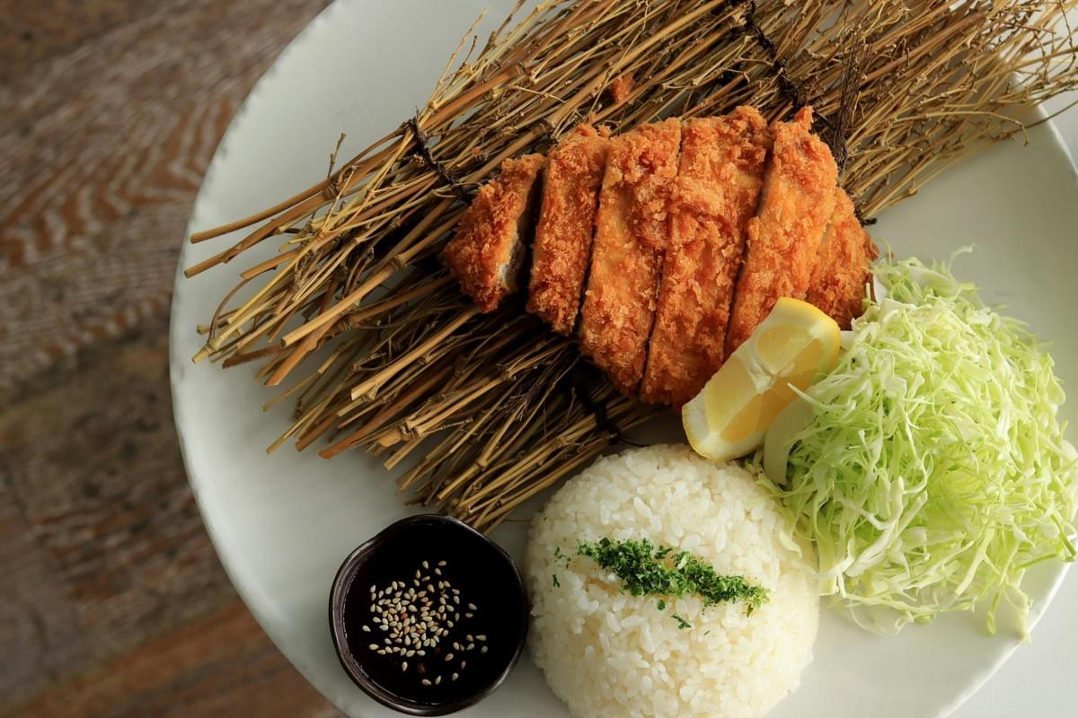 A high-angle shot features crispy, sliced tonkatsu served over a decorative bundle of straw alongside a scoop of white rice, shredded cabbage, and a fresh lemon wedge. A small side dish filled with dark dipping sauce and sprinkled with sesame seeds rests next to the rice on a large white plate. A high-angle shot features crispy, sliced tonkatsu served over a decorative bundle of straw alongside a scoop of white rice, shredded cabbage, and a fresh lemon wedge. A small side dish filled with dark dipping sauce and sprinkled with sesame seeds rests next to the rice on a large white plate.