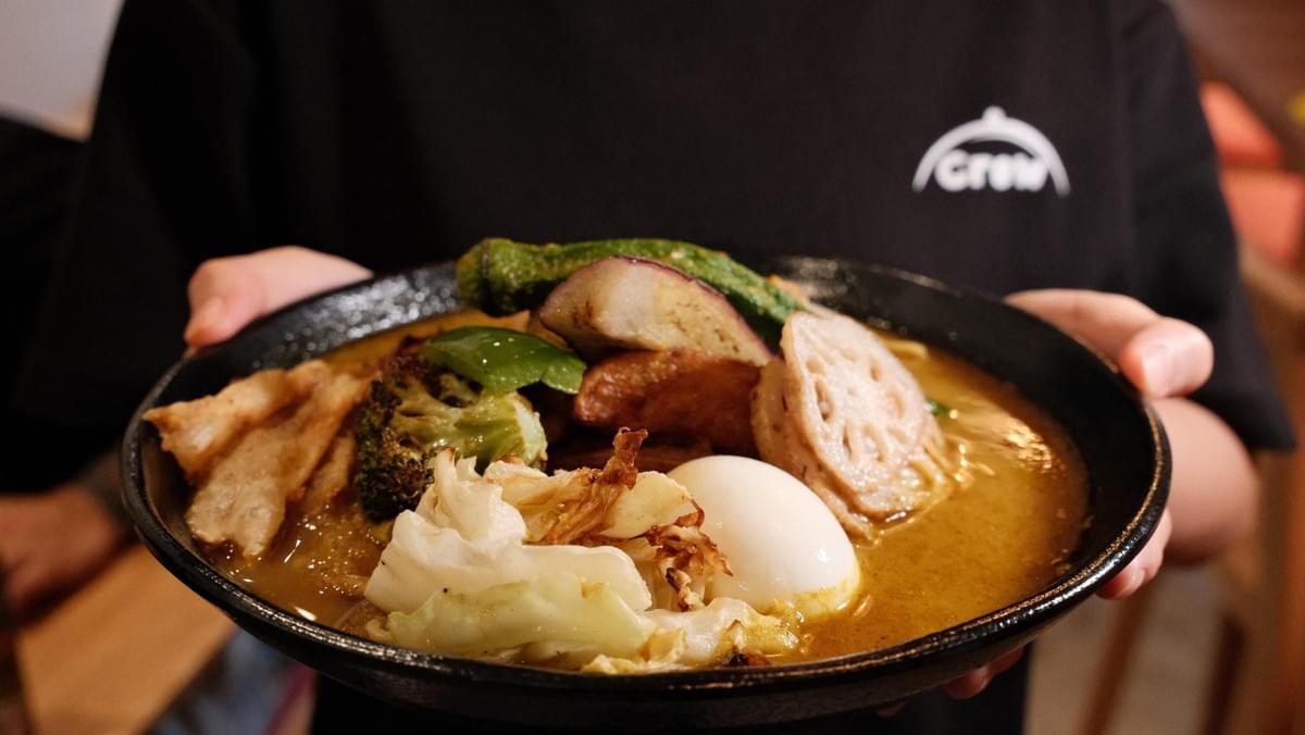 A staff member wearing a black t-shirt presents a wide black bowl of soup curry. The dish is rich with broth and topped with various ingredients, including a soft-boiled egg, lotus root, broccoli, cabbage, and eggplants. A staff member wearing a black t-shirt presents a wide black bowl of soup curry. The dish is rich with broth and topped with various ingredients, including a soft-boiled egg, lotus root, broccoli, cabbage, and eggplants.