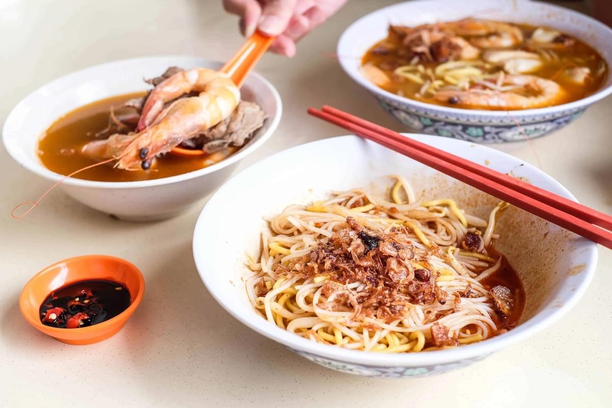 A large white bowl of noodles topped with fried shallots rests in the foreground with a pair of red chopsticks placed across its rim. In the background, a person scoops a cooked prawn out of a bowl of broth using an orange spoon. A large white bowl of noodles topped with fried shallots rests in the foreground with a pair of red chopsticks placed across its rim. In the background, a person scoops a cooked prawn out of a bowl of broth using an orange spoon.