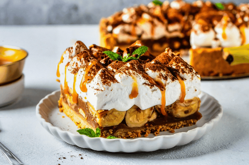 A decadent slice of banoffee pie on a white fluted plate, featuring a biscuit base, bananas, and a thick layer of whipped cream dusted with cocoa powder, drizzled with caramel, and garnished with a sprig of fresh mint. A decadent slice of banoffee pie on a white fluted plate, featuring a biscuit base, bananas, and a thick layer of whipped cream dusted with cocoa powder, drizzled with caramel, and garnished with a sprig of fresh mint.