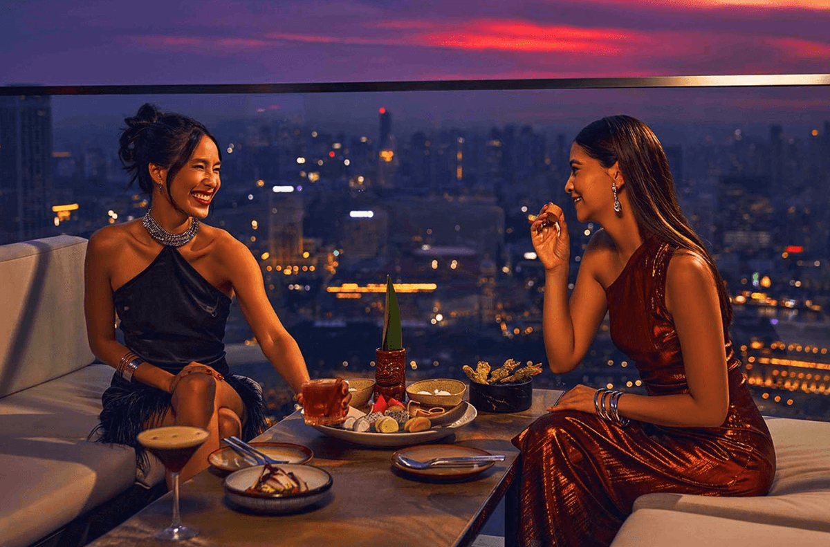 Two women elegantly dressed enjoy cocktails and snacks on a rooftop at sunset, with a vibrant cityscape in the background. They sit, smiling and laughing, creating a joyful and lively atmosphere. Two women elegantly dressed enjoy cocktails and snacks on a rooftop at sunset, with a vibrant cityscape in the background. They sit, smiling and laughing, creating a joyful and lively atmosphere.