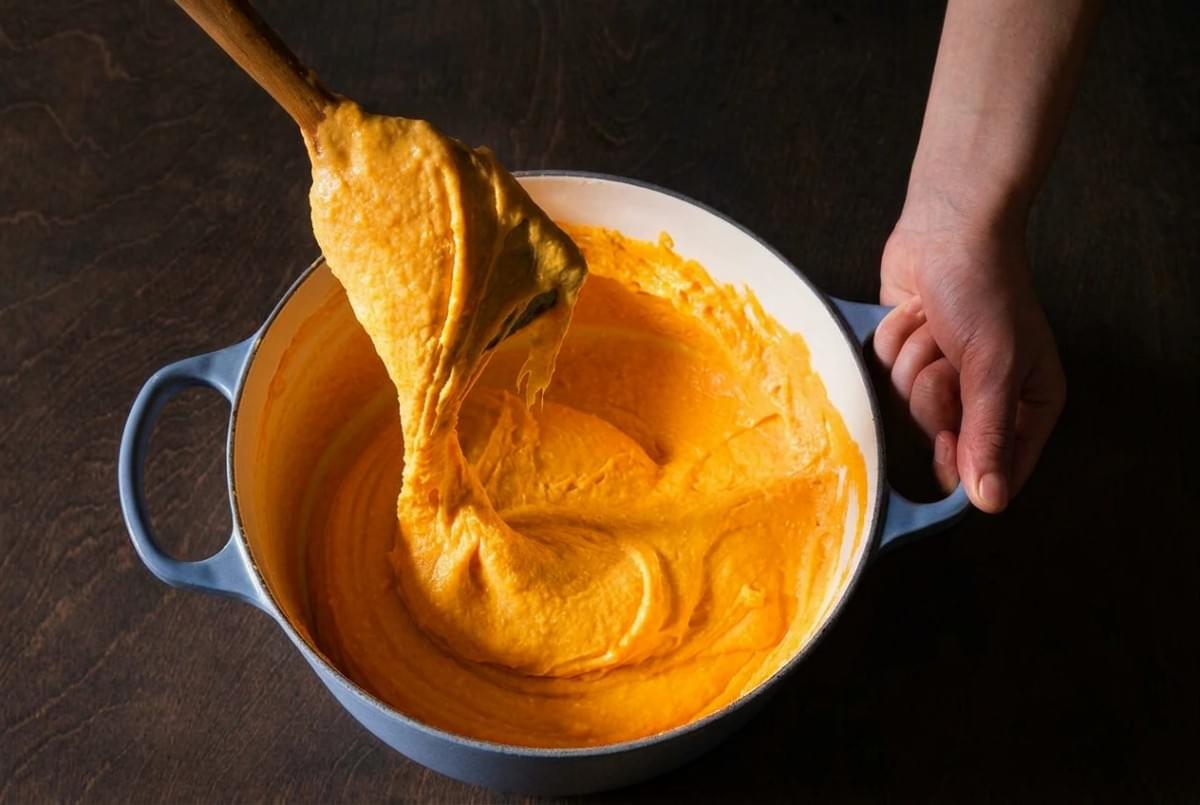 A wooden spoon lifting smooth, thick, bright orange sweet potato cheese sauce from a light blue Dutch oven. A wooden spoon lifting smooth, thick, bright orange sweet potato cheese sauce from a light blue Dutch oven.