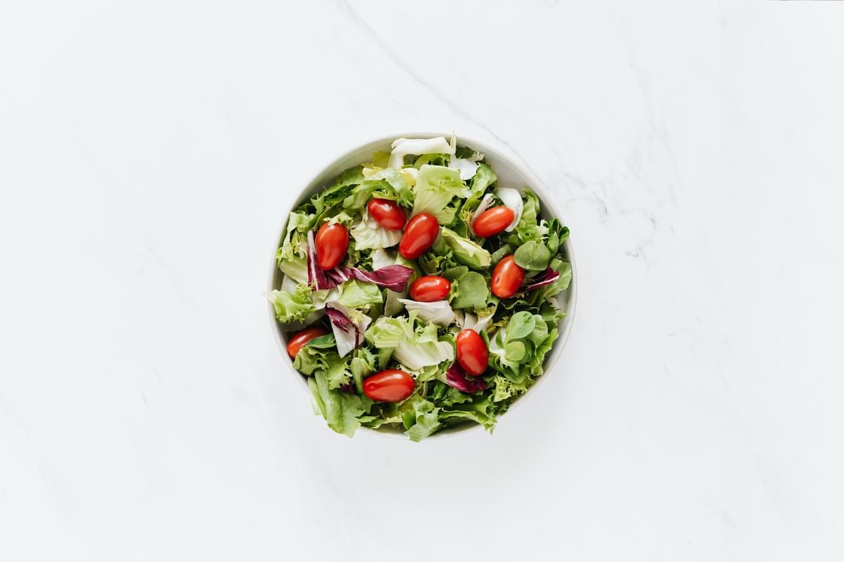 A top-down view shows a round white bowl filled with a fresh salad mix of green and purple lettuce leaves. Bright red cherry tomatoes are scattered across the top of the greens, all centered against a clean white marble background. A top-down view shows a round white bowl filled with a fresh salad mix of green and purple lettuce leaves. Bright red cherry tomatoes are scattered across the top of the greens, all centered against a clean white marble background.
