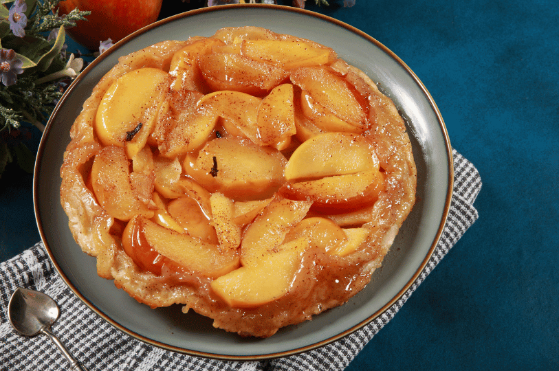 Top-down view of a traditional Tarte Tatin, an upside-down caramelized apple tart, resting on a dark plate against a dark blue background. Top-down view of a traditional Tarte Tatin, an upside-down caramelized apple tart, resting on a dark plate against a dark blue background.