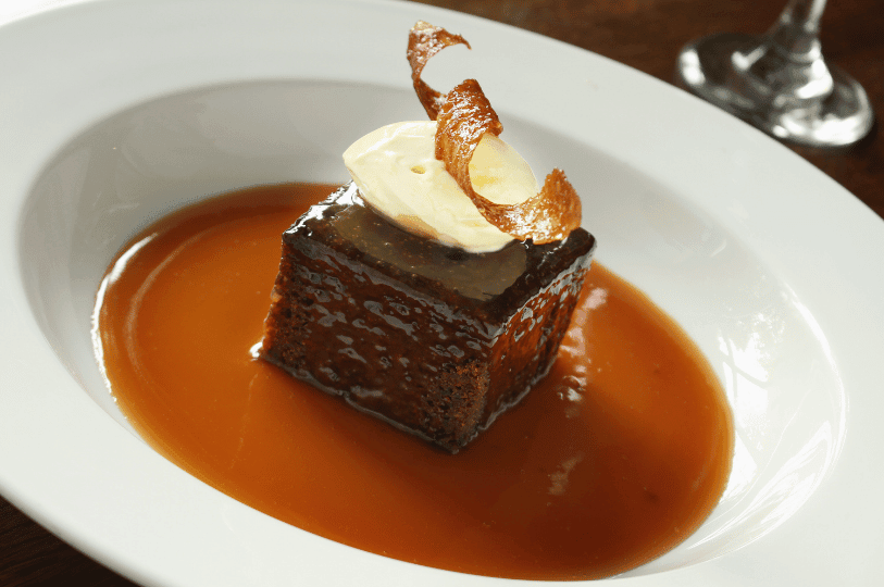 An elegantly plated square of sticky toffee pudding resting in a generous layer of caramel sauce, topped with a dollop of cream and a delicate, curled garnish. An elegantly plated square of sticky toffee pudding resting in a generous layer of caramel sauce, topped with a dollop of cream and a delicate, curled garnish.