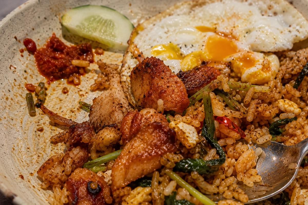 A top-down, close-up view shows a partially eaten plate of kampung fried rice mixed with seared chicken pieces, green leafy vegetables, and a fried egg with a runny yellow yolk. A metal spoon rests in the dish alongside a slice of cucumber and a smear of red chili sambal on the plate's rim. A top-down, close-up view shows a partially eaten plate of kampung fried rice mixed with seared chicken pieces, green leafy vegetables, and a fried egg with a runny yellow yolk. A metal spoon rests in the dish alongside a slice of cucumber and a smear of red chili sambal on the plate's rim.