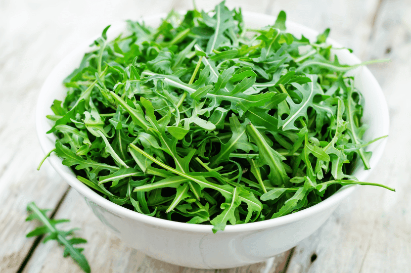 A bright white bowl generously overflowing with fresh spring arugula leaves on a light wooden surface. A bright white bowl generously overflowing with fresh spring arugula leaves on a light wooden surface.