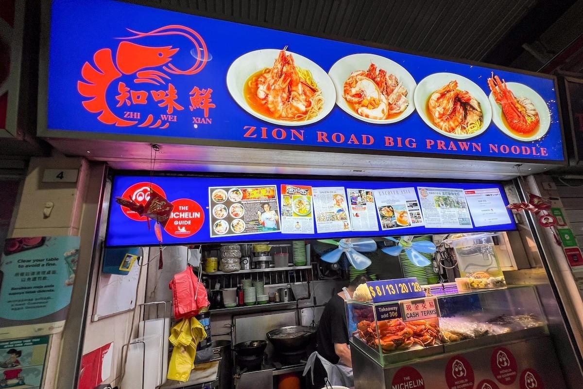 A illuminated blue sign reads "Zion Road Big Prawn Noodle" alongside a red prawn graphic, Chinese characters, and photos of dish options. Below the main sign, newspaper clippings and a Michelin Guide logo decorate the hawker stall counter. A illuminated blue sign reads "Zion Road Big Prawn Noodle" alongside a red prawn graphic, Chinese characters, and photos of dish options. Below the main sign, newspaper clippings and a Michelin Guide logo decorate the hawker stall counter.