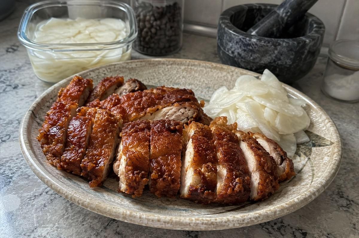 A high-angle, medium shot showcases a beautifully arranged plate of sliced Shoyu Koji fried chicken alongside a pile of thinly sliced pickled daikon radish on a neutral-toned speckled countertop. The chicken pieces are cut into thick, uniform slices, revealing an incredibly juicy, tender, light-pink interior and a deeply browned, caramelized, and ultra-crispy skin. In the background, a glass container holding more pickled radishes sits to the left, while a jar of peppercorns, a dark marble mortar and pestle, and a small glass jar of salt are softly blurred on the right side of the frame. A high-angle, medium shot showcases a beautifully arranged plate of sliced Shoyu Koji fried chicken alongside a pile of thinly sliced pickled daikon radish on a neutral-toned speckled countertop. The chicken pieces are cut into thick, uniform slices, revealing an incredibly juicy, tender, light-pink interior and a deeply browned, caramelized, and ultra-crispy skin. In the background, a glass container holding more pickled radishes sits to the left, while a jar of peppercorns, a dark marble mortar and pestle, and a small glass jar of salt are softly blurred on the right side of the frame.