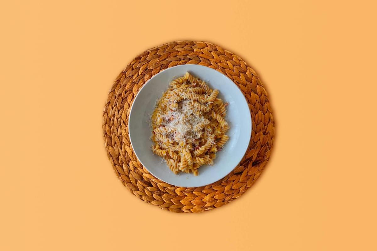 An overhead shot shows a plate of fusilli pasta tossed in a light orange sauce and topped with grated cheese, centered on a woven placemat. The entire arrangement is set against a solid, warm peach-colored background. An overhead shot shows a plate of fusilli pasta tossed in a light orange sauce and topped with grated cheese, centered on a woven placemat. The entire arrangement is set against a solid, warm peach-colored background.
