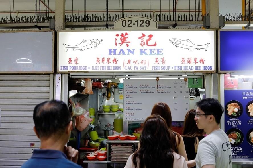 A group of customers queues in front of a food stall named Han Kee, unit 02-129, displaying a illuminated sign with fish illustrations and menu items. Below the sign, a menu board lists dish options and prices next to the kitchen preparation counter. A group of customers queues in front of a food stall named Han Kee, unit 02-129, displaying a illuminated sign with fish illustrations and menu items. Below the sign, a menu board lists dish options and prices next to the kitchen preparation counter.