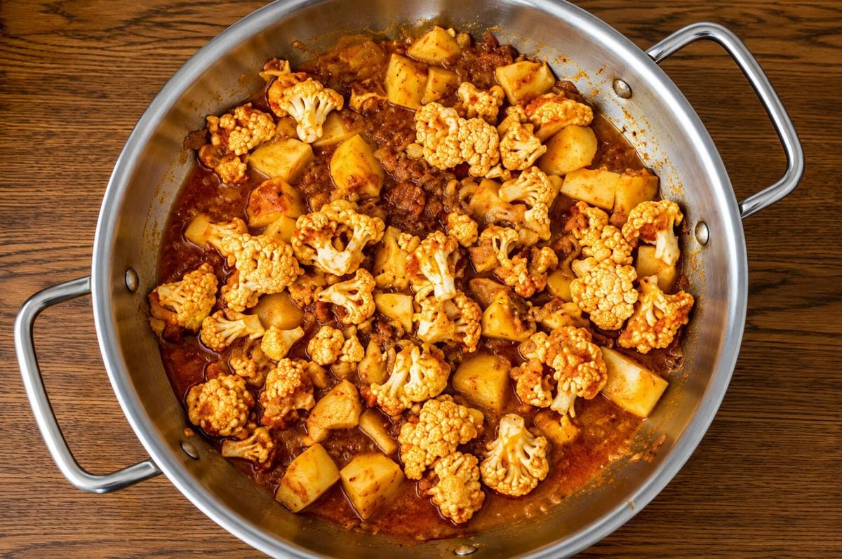 Overhead view of cauliflower florets and cubed potatoes simmering in a spiced tomato sauce inside a stainless steel skillet. Overhead view of cauliflower florets and cubed potatoes simmering in a spiced tomato sauce inside a stainless steel skillet.