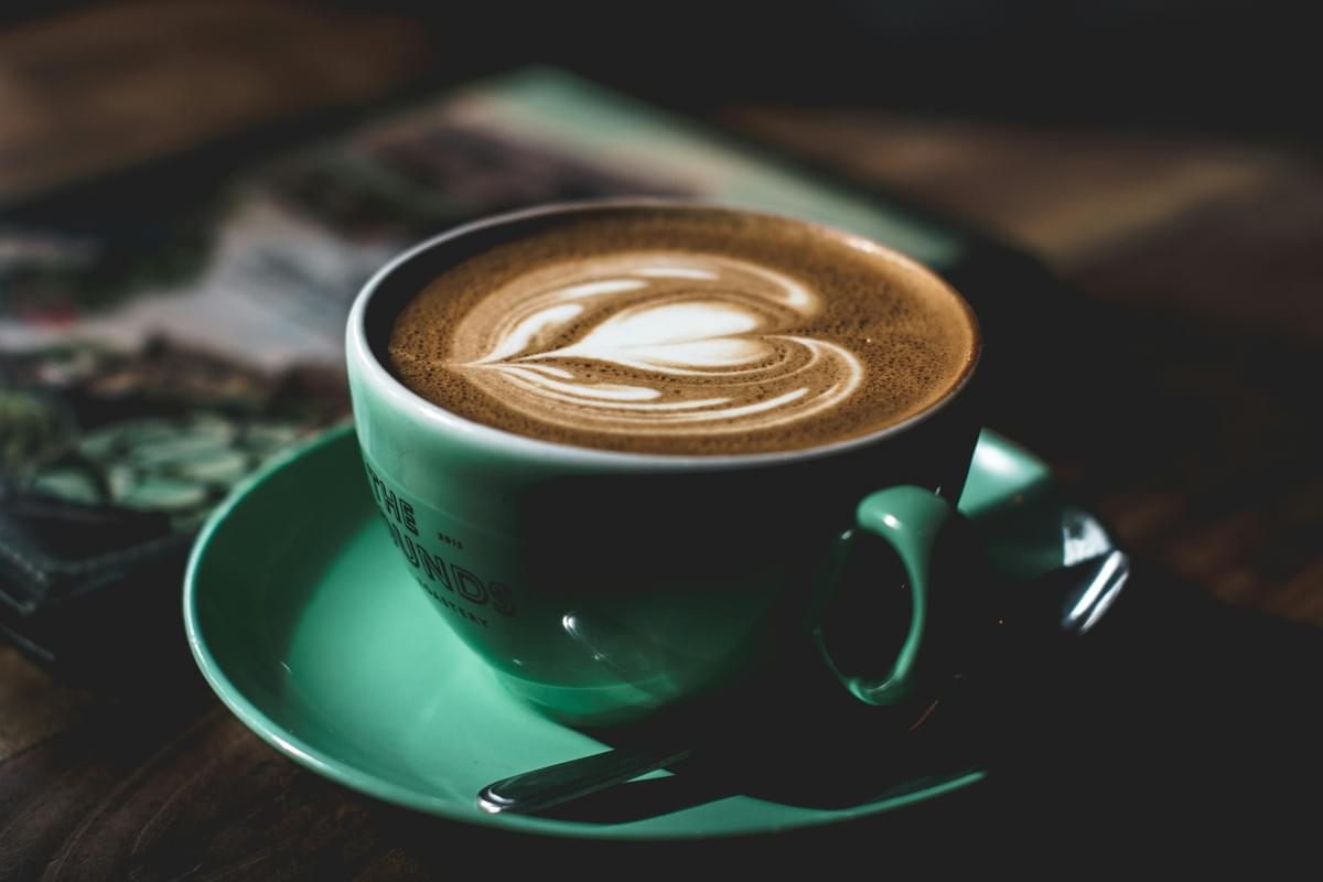 A teal ceramic cup filled with hot latte coffee sits on a matching teal saucer next to a small spoon. The coffee is adorned with a detailed heart-shaped latte art design made of microfoam. A teal ceramic cup filled with hot latte coffee sits on a matching teal saucer next to a small spoon. The coffee is adorned with a detailed heart-shaped latte art design made of microfoam.