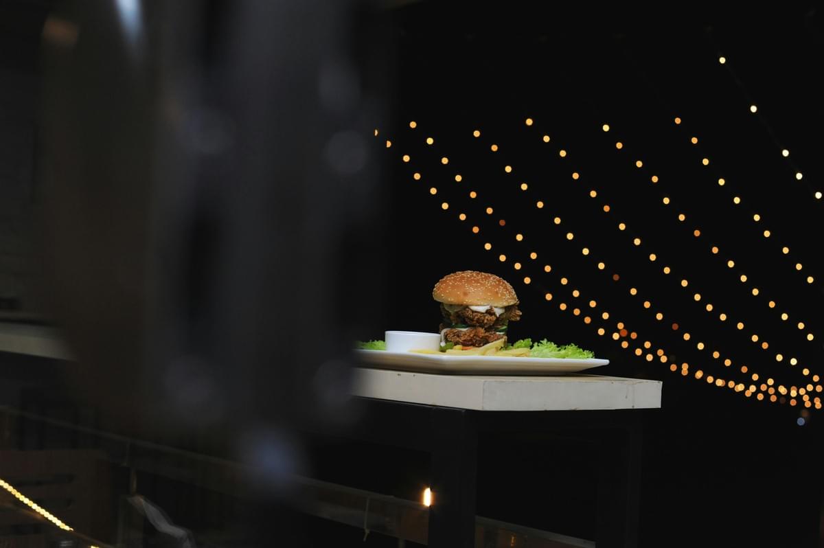 A double-decker fried chicken burger sits on a white plate alongside French fries and a side dipping sauce, placed on a high-top table. The background features a dark night setting illuminated by rows of warm, golden bokeh string lights, partially framed by a blurry dark object in the foreground. A double-decker fried chicken burger sits on a white plate alongside French fries and a side dipping sauce, placed on a high-top table. The background features a dark night setting illuminated by rows of warm, golden bokeh string lights, partially framed by a blurry dark object in the foreground.