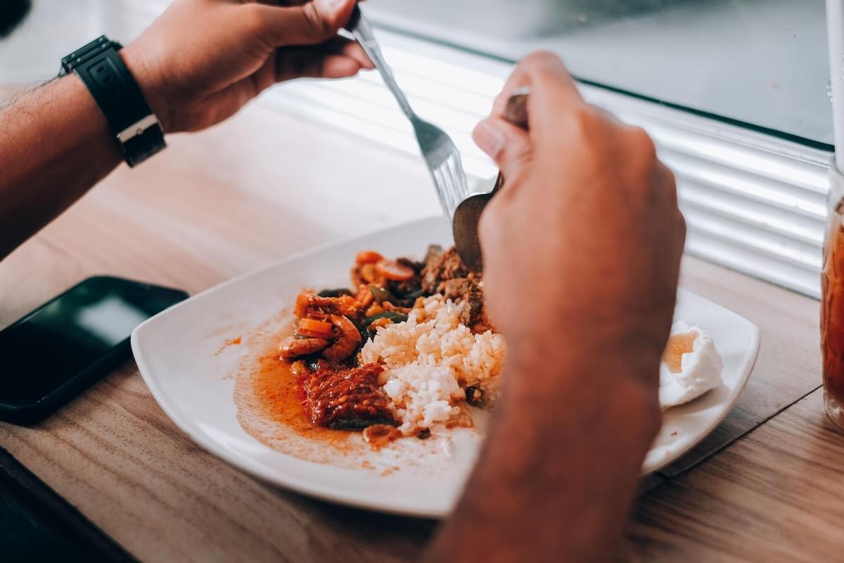 A close-up shot shows a person using a spoon and fork to eat a meal consisting of rice, meats, and vegetables served on a white plate. A smartphone rests face up on the wooden table nearby, next to the plate. A close-up shot shows a person using a spoon and fork to eat a meal consisting of rice, meats, and vegetables served on a white plate. A smartphone rests face up on the wooden table nearby, next to the plate.