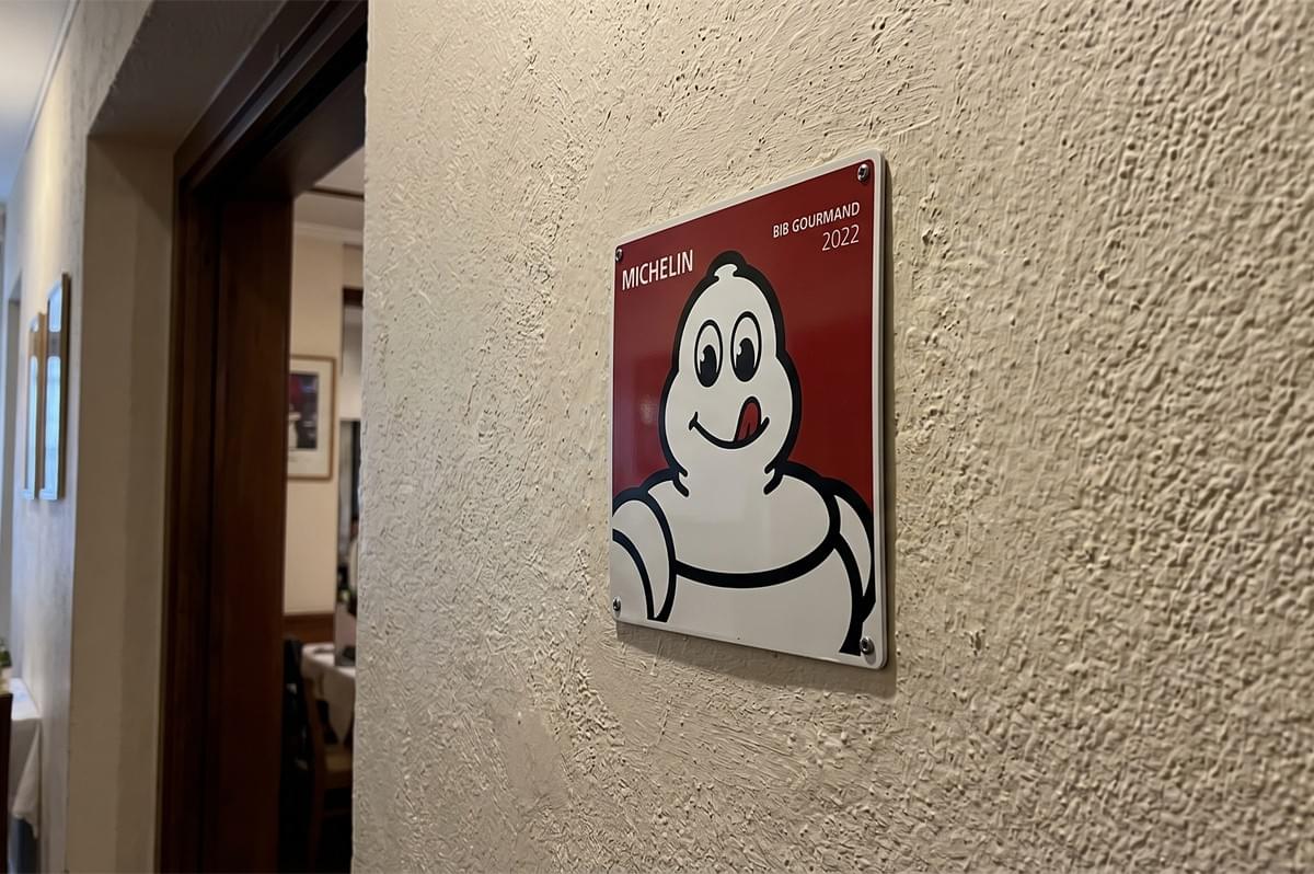 A Michelin Bib Gourmand 2022 award plaque featuring the Michelin Man mounted on an interior wall. A Michelin Bib Gourmand 2022 award plaque featuring the Michelin Man mounted on an interior wall.