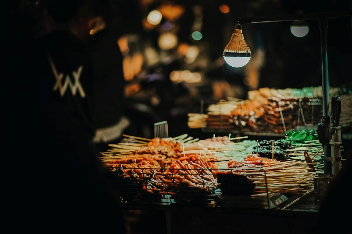 A vibrant night market stall displays an assortment of skewered street foods illuminated by a single, warm hanging bulb. The background is softly blurred, capturing the bustling atmosphere of a crowded outdoor evening scene. A vibrant night market stall displays an assortment of skewered street foods illuminated by a single, warm hanging bulb. The background is softly blurred, capturing the bustling atmosphere of a crowded outdoor evening scene.