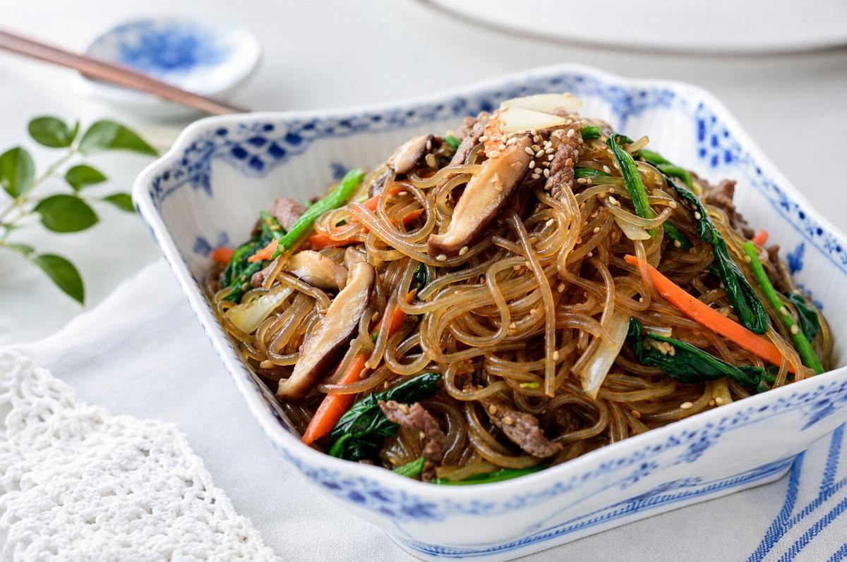 A blue-patterned ceramic bowl piled high with Japchae and mushrooms on a white tablecloth. A blue-patterned ceramic bowl piled high with Japchae and mushrooms on a white tablecloth.