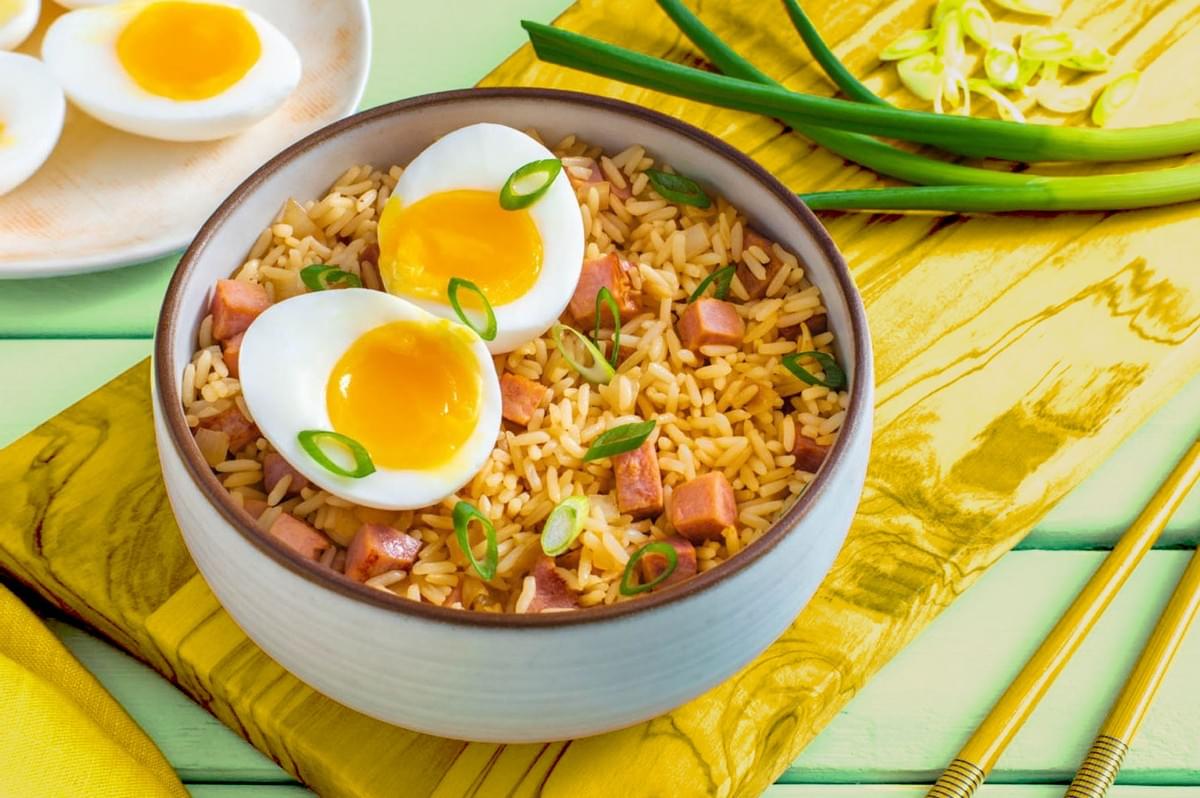 A bowl of fried rice with diced ham, garnished with sliced green onions and two soft-boiled eggs. A fresh and appetizing presentation, accompanied by a plate of halved eggs and whole green onions on the side. A bowl of fried rice with diced ham, garnished with sliced green onions and two soft-boiled eggs. A fresh and appetizing presentation, accompanied by a plate of halved eggs and whole green onions on the side.