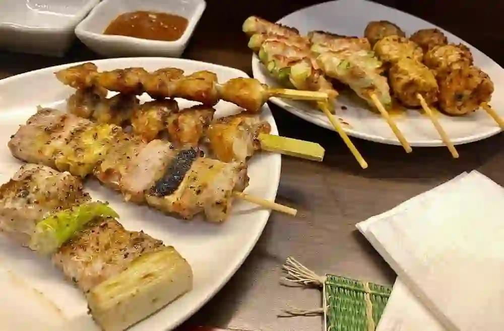 A variety of grilled yakitori skewers, including pork belly with leeks and chicken meatballs, are served on white plates at a restaurant table. A small dish of dipping sauce and a folded napkin sit nearby on the dark wooden surface. A variety of grilled yakitori skewers, including pork belly with leeks and chicken meatballs, are served on white plates at a restaurant table. A small dish of dipping sauce and a folded napkin sit nearby on the dark wooden surface.