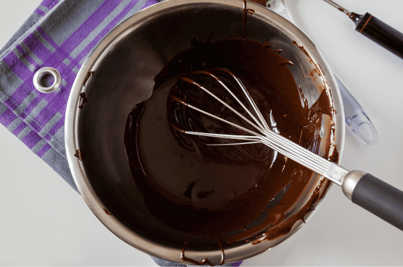 Top-down view of smooth, perfectly glossy tempered chocolate being stirred with a wire whisk inside a stainless steel mixing bowl resting on a purple and grey towel. Top-down view of smooth, perfectly glossy tempered chocolate being stirred with a wire whisk inside a stainless steel mixing bowl resting on a purple and grey towel.