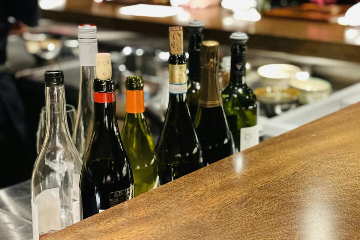 A variety of wine and sake bottles are lined up behind a polished wooden counter in a dimly lit bar or restaurant. The shallow depth of field keeps the bottles in sharp focus while the background kitchen area remains softly blurred. A variety of wine and sake bottles are lined up behind a polished wooden counter in a dimly lit bar or restaurant. The shallow depth of field keeps the bottles in sharp focus while the background kitchen area remains softly blurred.