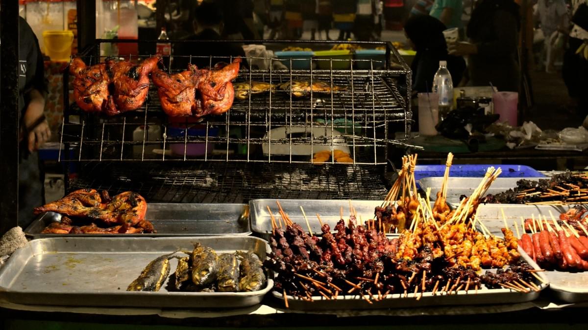 A vibrant street food stall displays an array of grilled delicacies, including whole charred chickens on a rack above trays of skewered meats, sausages, and grilled fish. A vibrant street food stall displays an array of grilled delicacies, including whole charred chickens on a rack above trays of skewered meats, sausages, and grilled fish.