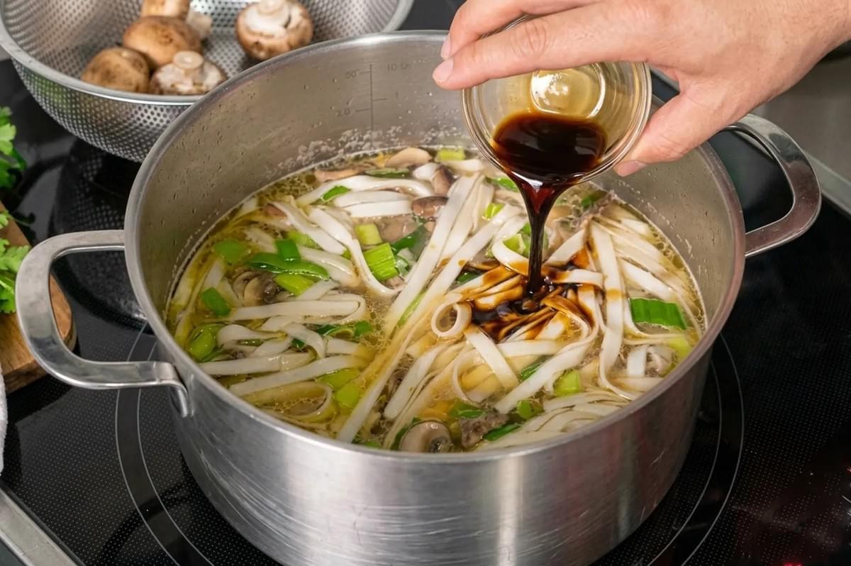 Hand pouring soy sauce into a large pot of boiling noodle soup with mushrooms and scallions Hand pouring soy sauce into a large pot of boiling noodle soup with mushrooms and scallions