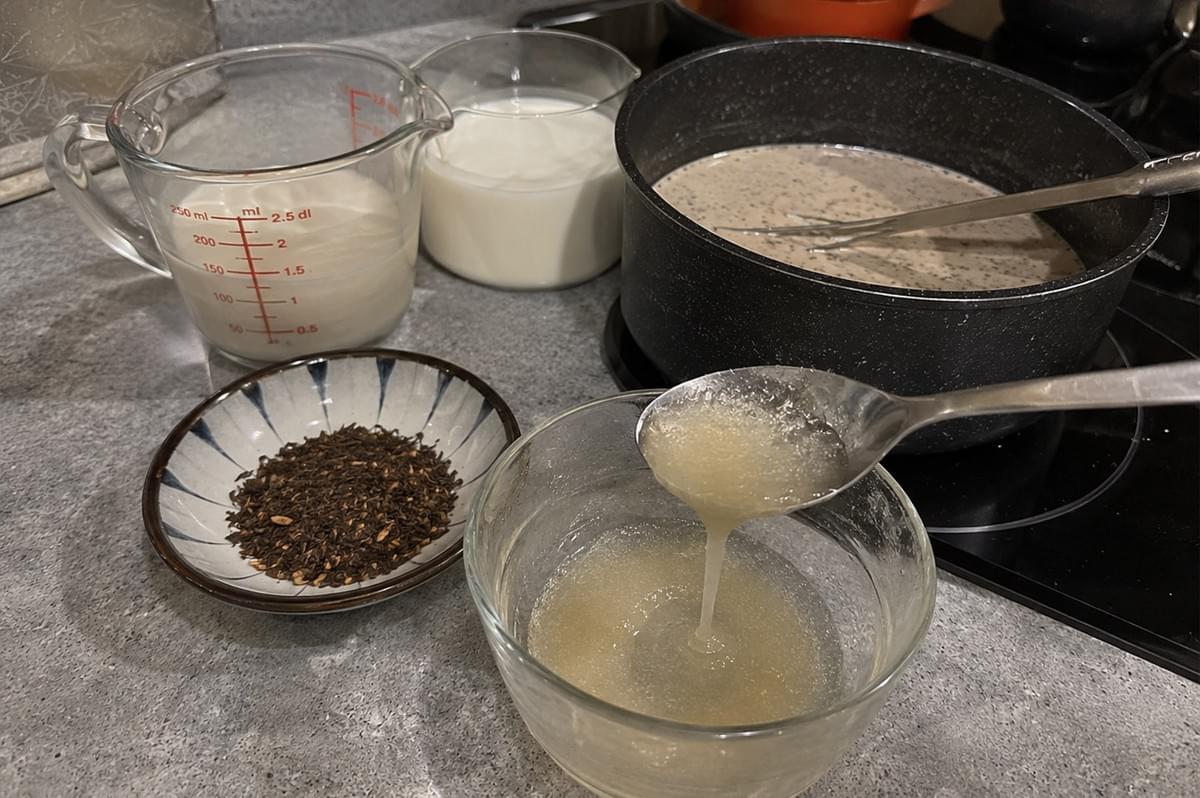 A kitchen countertop setup showing preparation steps, including glass measuring cups filled with cream and milk, a small ceramic dish containing loose hojicha tea leaves, a spoon dripping a clear gelled liquid over a glass bowl, and a pot with warm milk infused with tea on a cooktop. A kitchen countertop setup showing preparation steps, including glass measuring cups filled with cream and milk, a small ceramic dish containing loose hojicha tea leaves, a spoon dripping a clear gelled liquid over a glass bowl, and a pot with warm milk infused with tea on a cooktop.