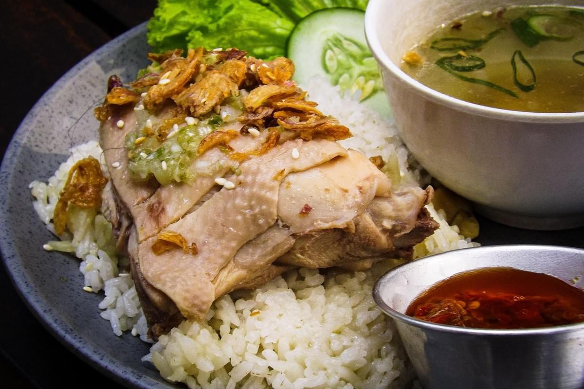 A close-up shot of a plate of Hainanese chicken rice featuring a piece of poached chicken topped with crispy fried shallots, green ginger sauce, and sesame seeds served over a bed of seasoned rice. The dish is accompanied by a bowl of clear broth with chopped scallions, a small metal cup of red chili dipping sauce, a slice of cucumber, and lettuce. A close-up shot of a plate of Hainanese chicken rice featuring a piece of poached chicken topped with crispy fried shallots, green ginger sauce, and sesame seeds served over a bed of seasoned rice. The dish is accompanied by a bowl of clear broth with chopped scallions, a small metal cup of red chili dipping sauce, a slice of cucumber, and lettuce.