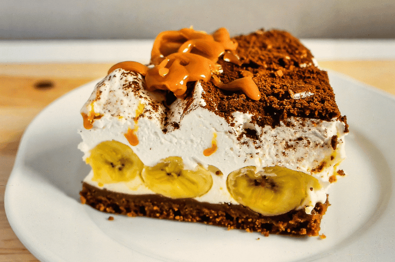 A square slice of banoffee pie on a white plate, displaying a dark, crumbly crust, sliced bananas, a thick layer of cream heavily dusted with cocoa powder, and thick caramel squiggles on top. A square slice of banoffee pie on a white plate, displaying a dark, crumbly crust, sliced bananas, a thick layer of cream heavily dusted with cocoa powder, and thick caramel squiggles on top.