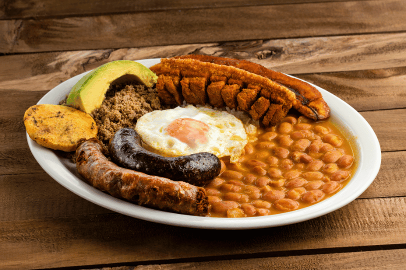 A traditional Colombian platter on a wooden surface, showcasing creamy beans, a sunny-side-up egg, morcilla (blood sausage), chorizo, ground meat, a slice of avocado, and a thick piece of crunchy chicharrón. A traditional Colombian platter on a wooden surface, showcasing creamy beans, a sunny-side-up egg, morcilla (blood sausage), chorizo, ground meat, a slice of avocado, and a thick piece of crunchy chicharrón.