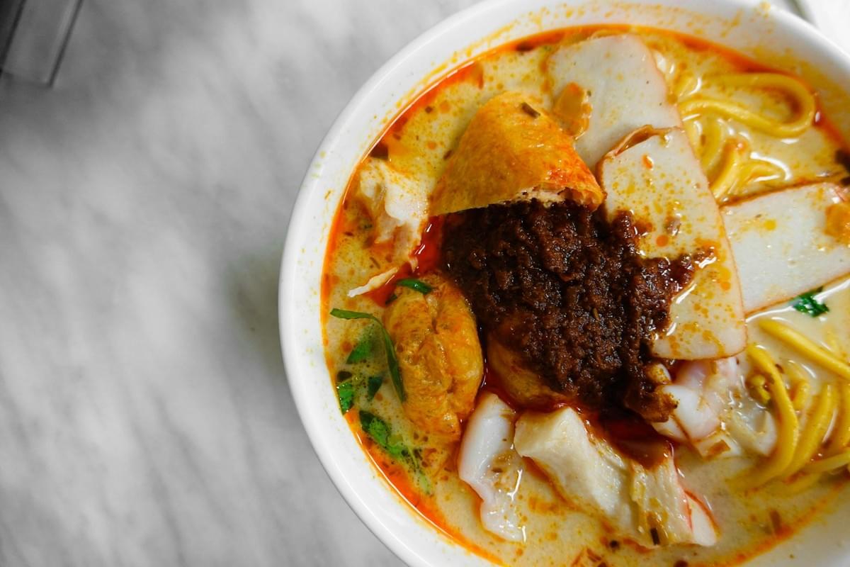 A white bowl sits on a light marble surface, filled with a rich laksa noodle soup in a spicy coconut broth. The dish is topped with yellow noodles, sliced fish cakes, tofu puffs, and a dark chili paste paste in the center. A white bowl sits on a light marble surface, filled with a rich laksa noodle soup in a spicy coconut broth. The dish is topped with yellow noodles, sliced fish cakes, tofu puffs, and a dark chili paste paste in the center.