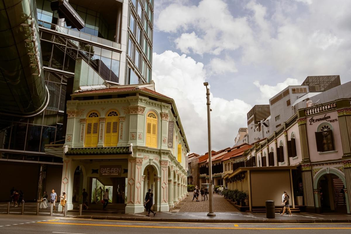 This image captures a striking contrast between the ornate, pastel-colored Peranakan-style shophouses and a towering, modern glass skyscraper in an urban setting. Pedestrians walk along the street level, where the historic architecture's intricate details and yellow shutters stand out against the bright, clouded sky. This image captures a striking contrast between the ornate, pastel-colored Peranakan-style shophouses and a towering, modern glass skyscraper in an urban setting. Pedestrians walk along the street level, where the historic architecture's intricate details and yellow shutters stand out against the bright, clouded sky.