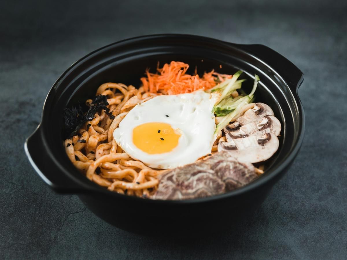 A black ceramic bowl filled with thick noodles and topped with a runny fried egg sits on a dark gray surface. The dish is neatly garnished with sliced mushrooms, shredded carrots, cucumber strips, meat, and seaweed flakes. A black ceramic bowl filled with thick noodles and topped with a runny fried egg sits on a dark gray surface. The dish is neatly garnished with sliced mushrooms, shredded carrots, cucumber strips, meat, and seaweed flakes.