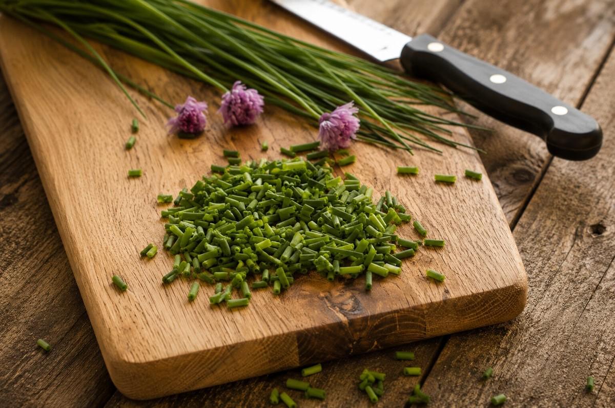 A pile of chopped chives next to whole chive stalks, purple chive blossoms, and a chef's knife on a wooden board. A pile of chopped chives next to whole chive stalks, purple chive blossoms, and a chef's knife on a wooden board.