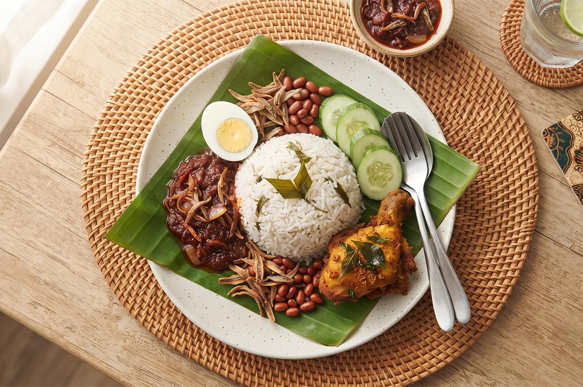 Top-down view of a plate of Nasi Lemak with coconut rice, fried chicken, sambal, peanuts, anchovies, boiled egg, and cucumber slices served on a banana leaf. Top-down view of a plate of Nasi Lemak with coconut rice, fried chicken, sambal, peanuts, anchovies, boiled egg, and cucumber slices served on a banana leaf.