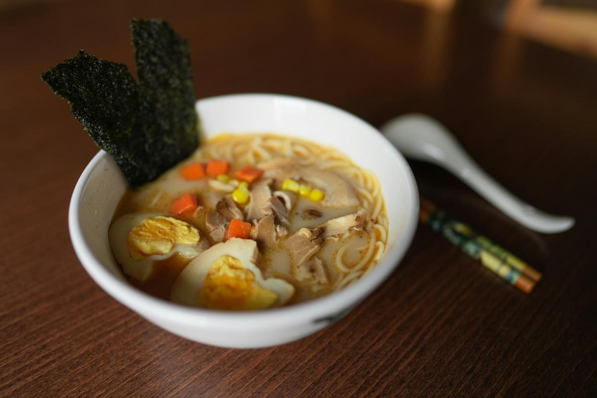 A steaming white bowl of ramen sits on a wooden table, filled with noodles, broth, sliced meat, corn, diced carrots, two soft-boiled egg halves, and a crisp sheet of nori seaweed. In the softly blurred background, a white soup spoon and a pair of chopsticks lie beside the bowl. A steaming white bowl of ramen sits on a wooden table, filled with noodles, broth, sliced meat, corn, diced carrots, two soft-boiled egg halves, and a crisp sheet of nori seaweed. In the softly blurred background, a white soup spoon and a pair of chopsticks lie beside the bowl.