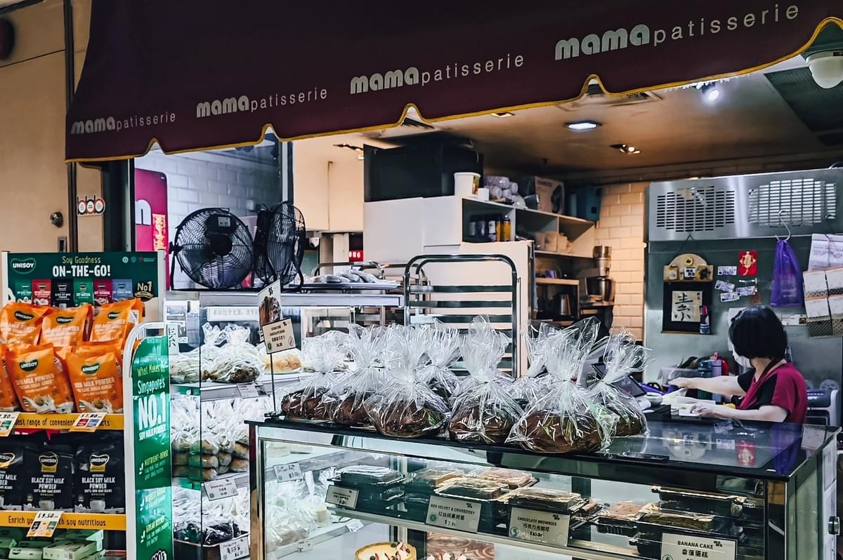 “Eye-level medium-wide shot of a Mama Patisserie bakery stall inside a mall food center, featuring a maroon awning with repeated ‘mama patisserie’ text, glass display cases filled with pastries and packaged baked goods, plastic-wrapped items lined on top, small electric fans, and a staff member working behind the counter under warm indoor lighting. “Eye-level medium-wide shot of a Mama Patisserie bakery stall inside a mall food center, featuring a maroon awning with repeated ‘mama patisserie’ text, glass display cases filled with pastries and packaged baked goods, plastic-wrapped items lined on top, small electric fans, and a staff member working behind the counter under warm indoor lighting.