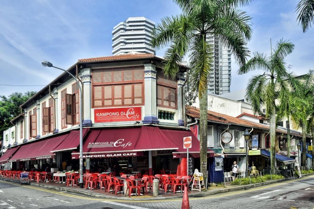 This image captures the Kampong Glam Cafe, a corner eatery featuring a prominent red awning and matching red outdoor seating in a historic shophouse. Tall palm trees and modern skyscrapers stand in the background, highlighting the contrast between the area's traditional architecture and the city's urban skyline. This image captures the Kampong Glam Cafe, a corner eatery featuring a prominent red awning and matching red outdoor seating in a historic shophouse. Tall palm trees and modern skyscrapers stand in the background, highlighting the contrast between the area's traditional architecture and the city's urban skyline.