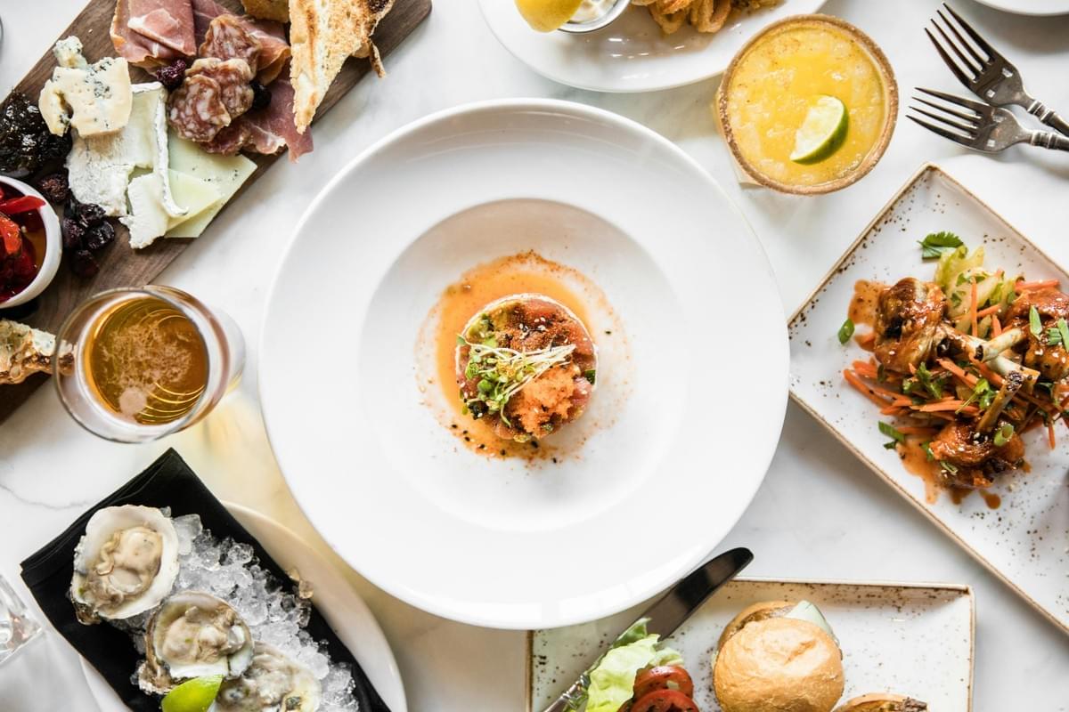 An overhead shot shows a marble tabletop spread with a variety of gourmet dishes, centered around a white bowl with a neatly stacked tuna tartare garnished with microgreens and roe. Surrounding the main plate are oysters on ice, a charcuterie board, glazed chicken wings, sliders, and refreshing beverages. An overhead shot shows a marble tabletop spread with a variety of gourmet dishes, centered around a white bowl with a neatly stacked tuna tartare garnished with microgreens and roe. Surrounding the main plate are oysters on ice, a charcuterie board, glazed chicken wings, sliders, and refreshing beverages.