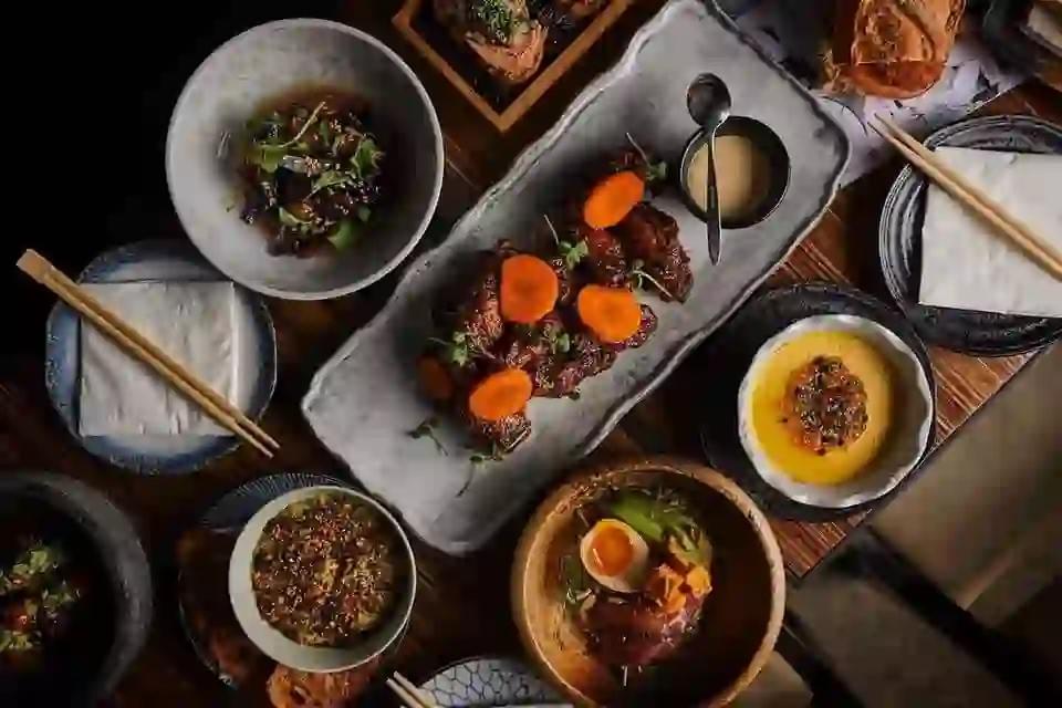 An overhead shot displays an assortment of Japanese-inspired fusion dishes, including glazed ribs with carrot slices and a soft-boiled egg bowl, arranged on a dark wooden table. Chopsticks, small dipping sauces, and textured ceramic plates complete the spread, creating an inviting and sophisticated dining atmosphere. An overhead shot displays an assortment of Japanese-inspired fusion dishes, including glazed ribs with carrot slices and a soft-boiled egg bowl, arranged on a dark wooden table. Chopsticks, small dipping sauces, and textured ceramic plates complete the spread, creating an inviting and sophisticated dining atmosphere.
