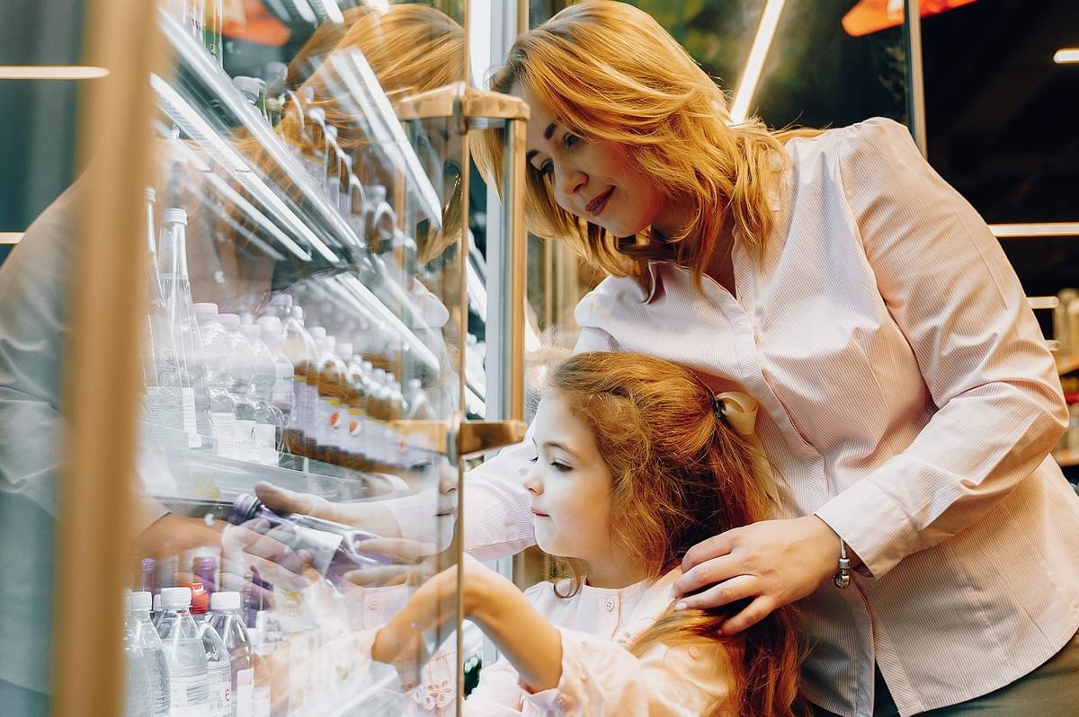 Over-the-shoulder medium shot of adult guiding child selecting drinks from a supermarket fridge, highlighting language learning, everyday grocery shopping abroad, and educational family experiences in foreign markets Over-the-shoulder medium shot of adult guiding child selecting drinks from a supermarket fridge, highlighting language learning, everyday grocery shopping abroad, and educational family experiences in foreign markets