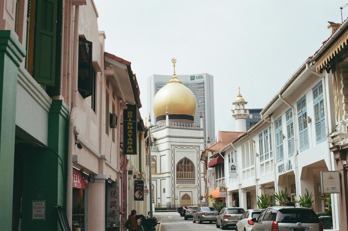A narrow city street is lined with colorful historic shophouses and parked cars. At the end of the street stands the grand Sultan Mosque, featuring a striking golden dome. A narrow city street is lined with colorful historic shophouses and parked cars. At the end of the street stands the grand Sultan Mosque, featuring a striking golden dome.