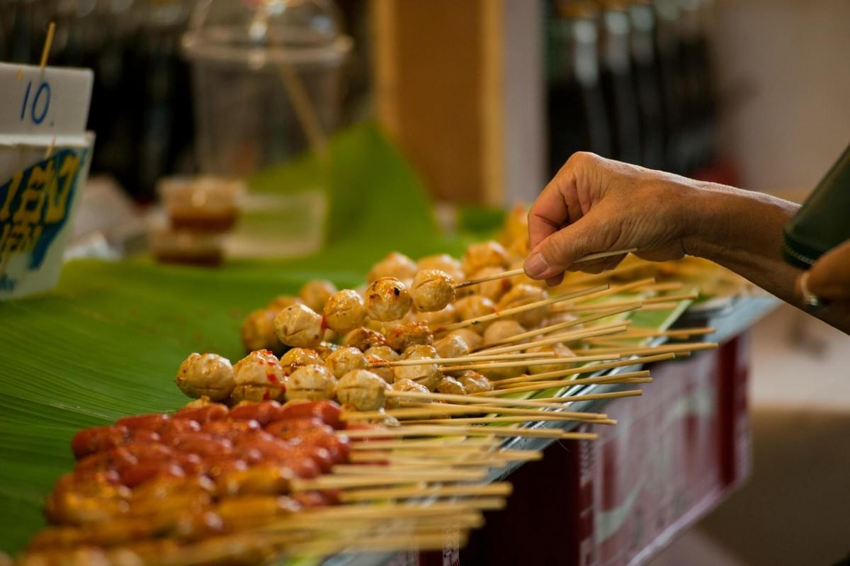 A person's hand reaches for one of many skewers of grilled meatballs and sausages arranged on bright green banana leaves at a vibrant street food stall. The shallow depth of field captures the bustling atmosphere of the market, highlighting the variety of skewers available for sale. A person's hand reaches for one of many skewers of grilled meatballs and sausages arranged on bright green banana leaves at a vibrant street food stall. The shallow depth of field captures the bustling atmosphere of the market, highlighting the variety of skewers available for sale.