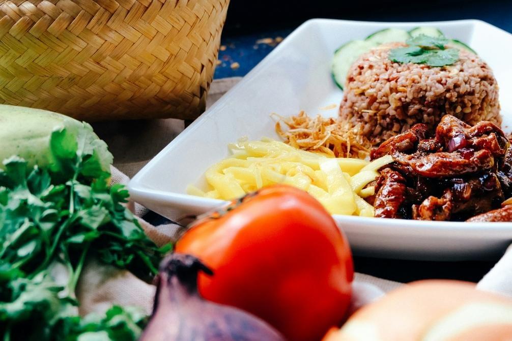 The image features a close-up shot of a white plate filled with Thai shrimp paste fried rice, sweet pork, shredded green mango, and sliced cucumbers. In the foreground, fresh ingredients like a ripe red tomato, onions, and cilantro are softly blurred next to a woven bamboo basket. The image features a close-up shot of a white plate filled with Thai shrimp paste fried rice, sweet pork, shredded green mango, and sliced cucumbers. In the foreground, fresh ingredients like a ripe red tomato, onions, and cilantro are softly blurred next to a woven bamboo basket.